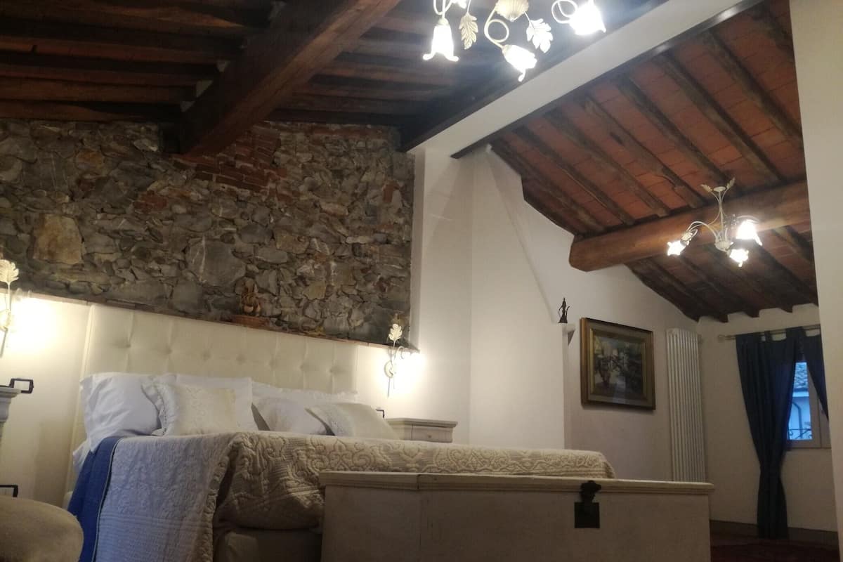 A bedroom with a stone wall, wooden beams, a bed with a white headboard, and a chandelier.