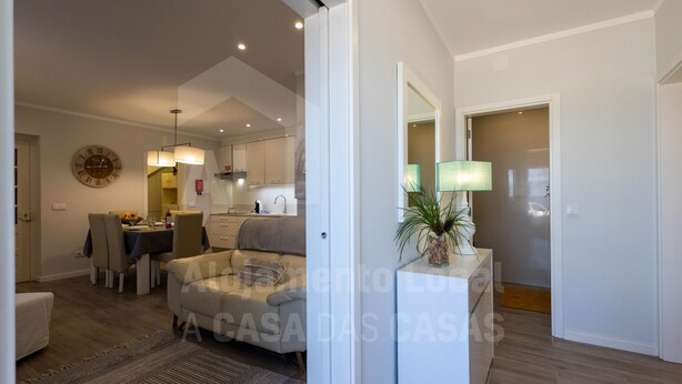 A modern living room with a sofa, a dining area with chairs, a kitchen in the background, and a hallway leading to another room.