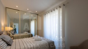 A hotel room with a large bed, a chair, a mirror, and a view of the street through sheer curtains.