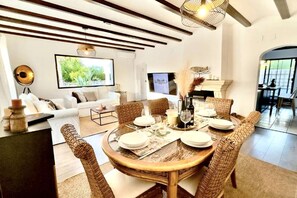 Un coin repas avec une table ronde dressée pour quatre personnes, entourée de chaises en osier. Un téléviseur est fixé au mur, et un canapé est visible au fond.