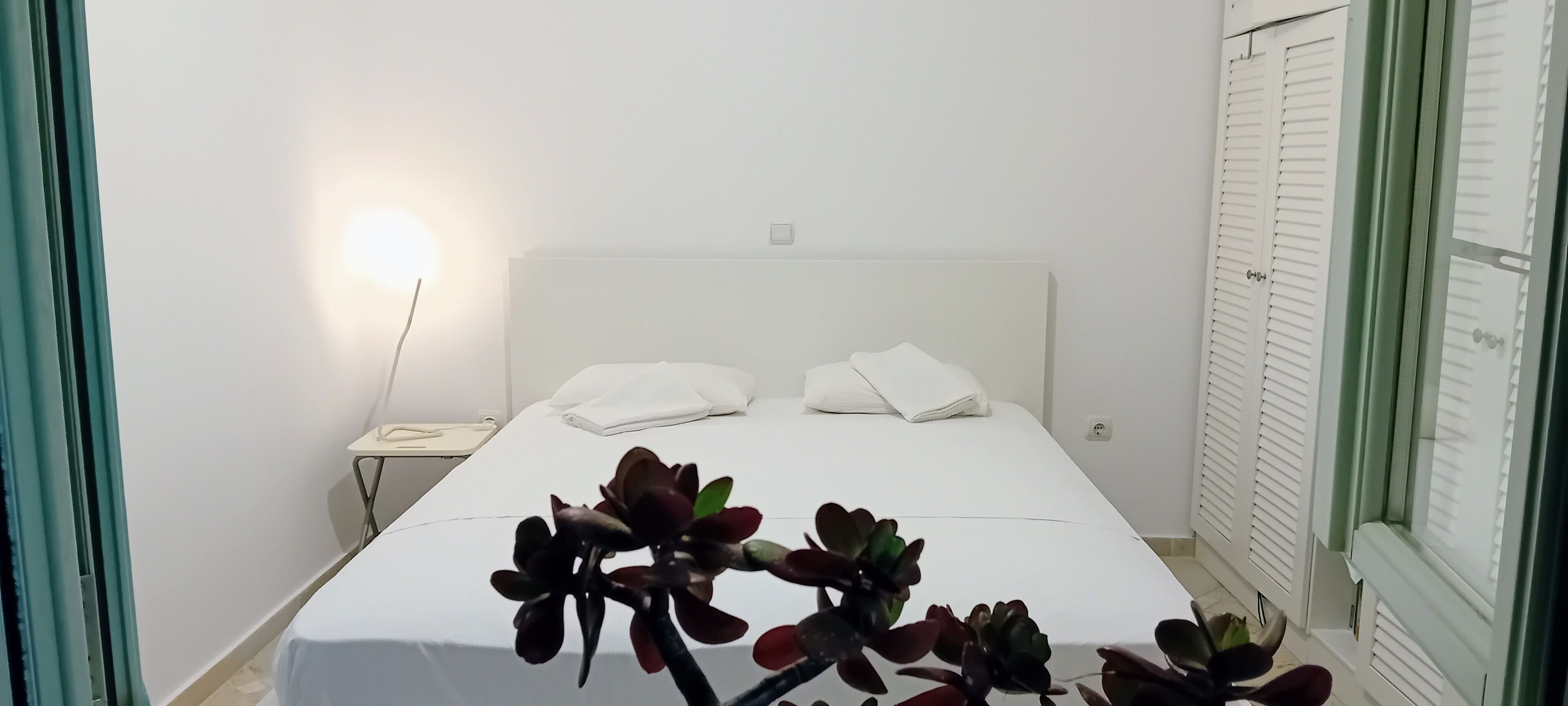 A neatly made bed with white linens, a bedside table with a lamp, and a window with blinds.