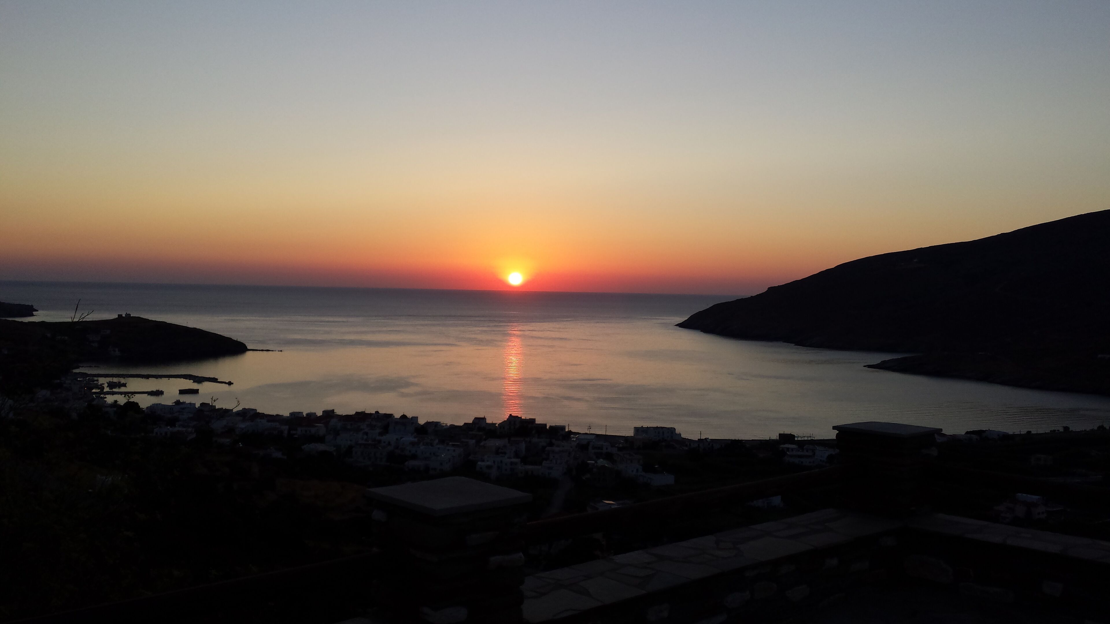 Vista de un atardecer costero con aguas tranquilas y el silueta de un pueblo al frente.