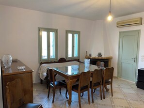 Un coin repas avec une table et des chaises en bois, deux fenêtres aux cadres verts, une cheminée et un appareil de climatisation.