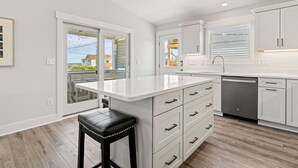 A modern kitchen with white cabinetry, a black bar stool, and a large window overlooking a residential area. - One More Moon 3 Bedroom Home in Kitty Hawk (Kitty Hawk)