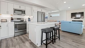 A modern kitchen with white cabinets, stainless steel appliances, a central island, and a blue sofa in the living area. - One More Moon 3 Bedroom Home in Kitty Hawk (Kitty Hawk)