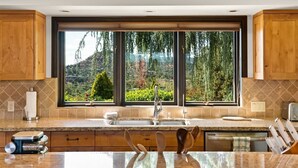 A kitchen with wooden cabinets, a granite countertop, a large window with a view of greenery, and a built-in dishwasher.