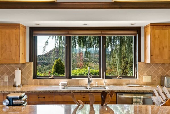 A kitchen with wooden cabinets, a granite countertop, a large window with a view of greenery, and a built-in dishwasher.