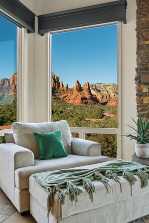 A room with a large window overlooking a mountainous landscape, a sofa with a green pillow, and a potted plant. - Luxury Paradise, 360 Views, Pool/Hot Tub/Firepit! (Sedona)