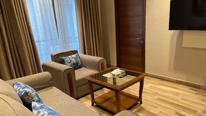 A living room with a sofa, armchairs, a glass coffee table, a wooden door, and a TV mounted on the wall. - UDS Villa - Next to VFS, Walking to Connaught Place (New Delhi)