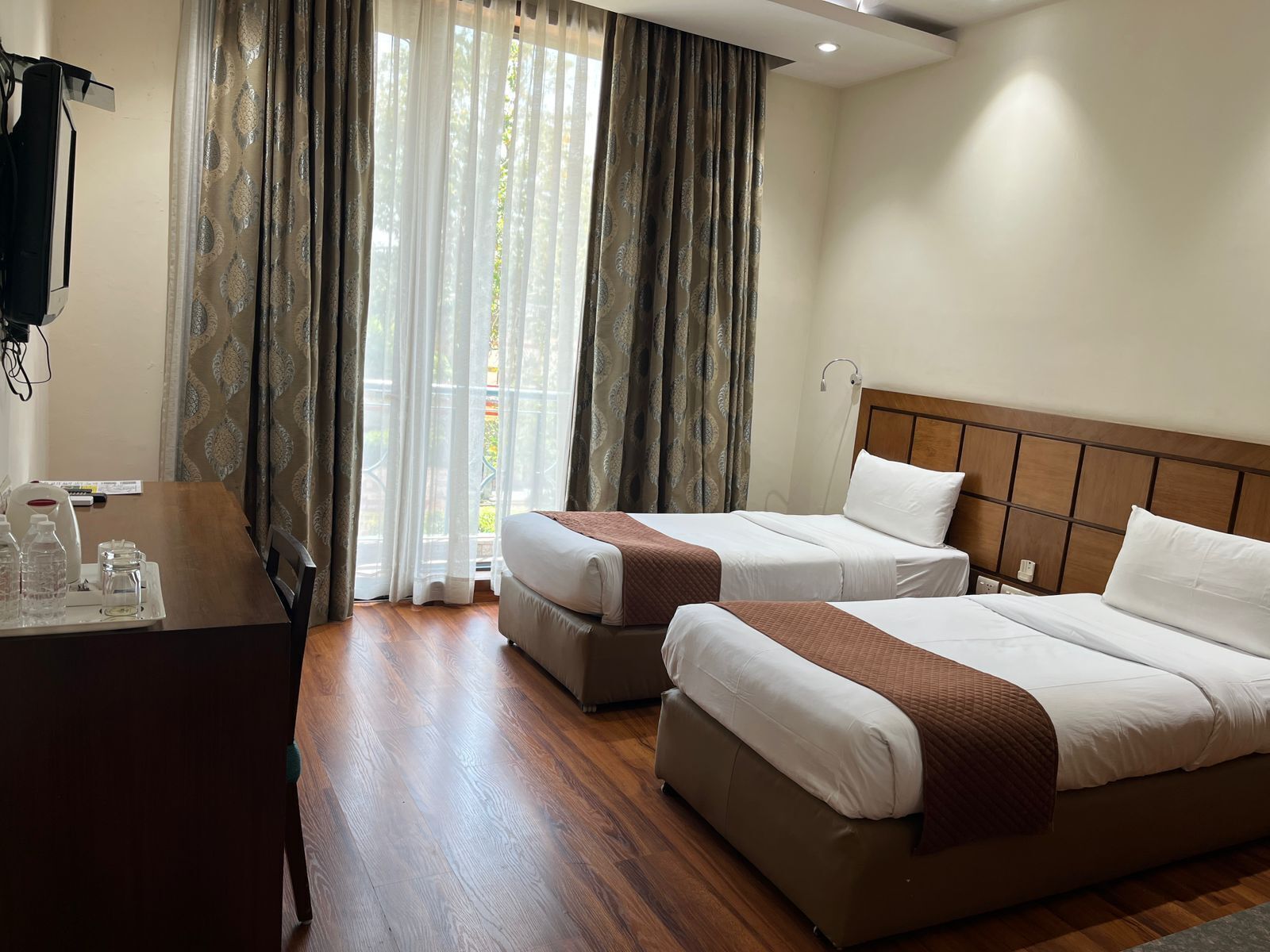 Quarto de hotel com duas camas, uma escrivaninha e vista através de cortinas leves.