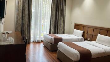 Quarto de hotel com duas camas, uma escrivaninha e vista através de cortinas leves.