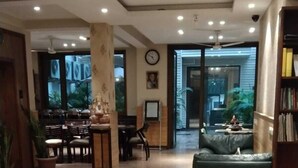A spacious lobby with a high ceiling, a seating area with a sofa and chairs, a dining area with tables and chairs, and a large window providing a view of the outdoors. - UDS Villa - Next to VFS, Walking to Connaught Place (New Delhi)