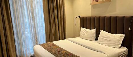 A hotel room with a large bed, a headboard, a bedside table, a lamp, and a window with curtains.
