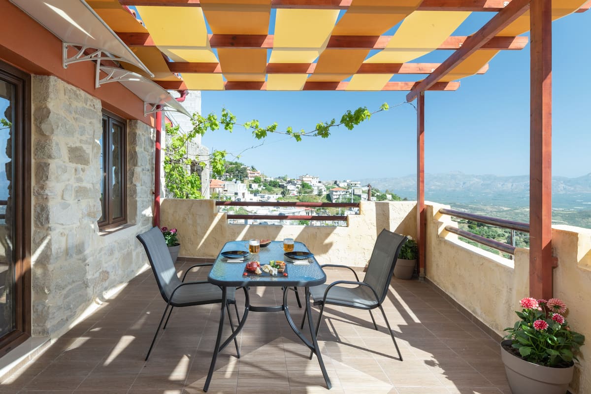 Een terras met een gedekte tafel voor twee, een pergola erboven en uitzicht op een heuvelstad.