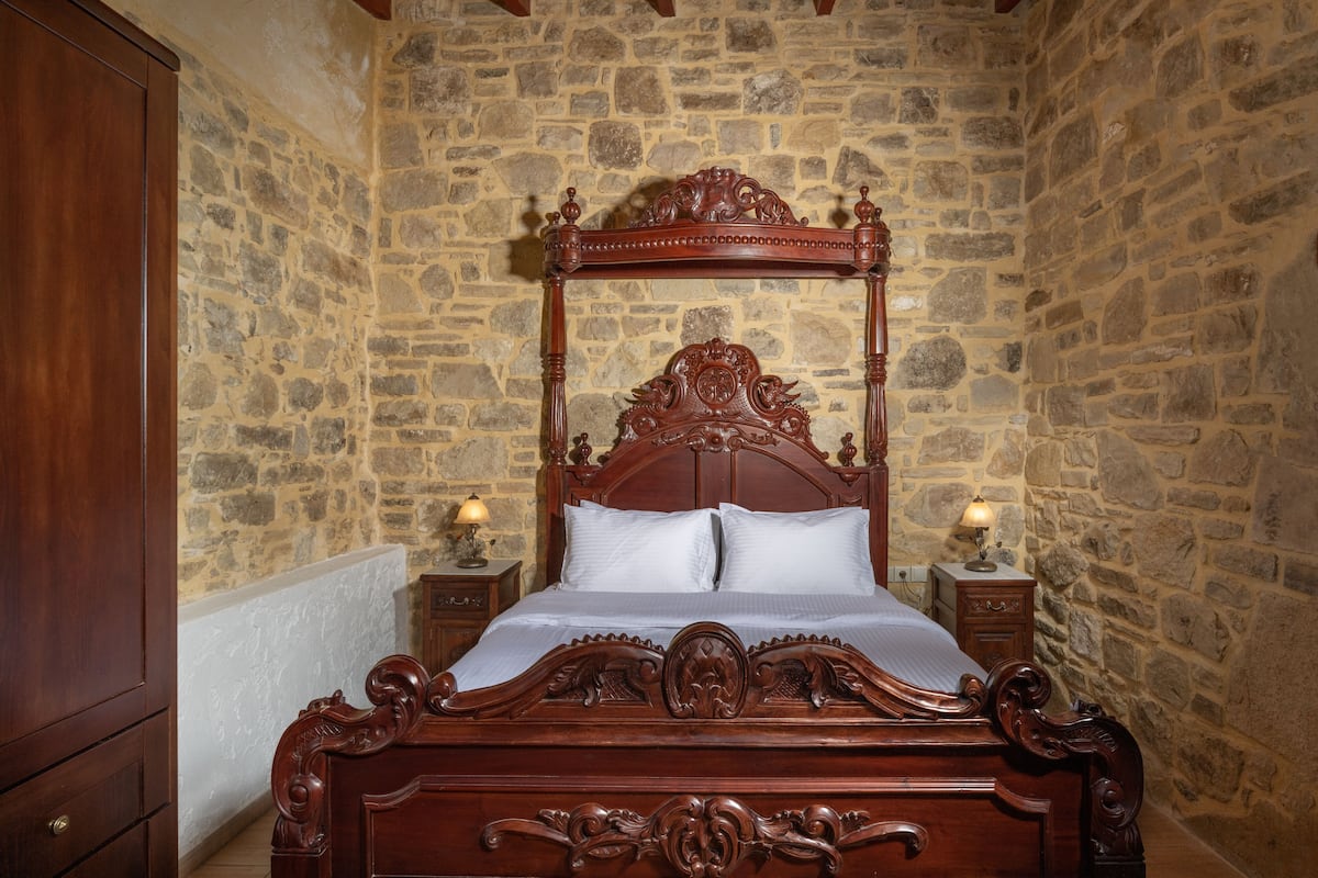 Een houten hemelbed met witte lakens in een kamer met stenen muren en houten meubels.