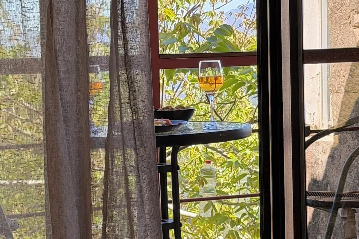 Een raam met uitzicht op een boom en een tafel met een glas wijn.