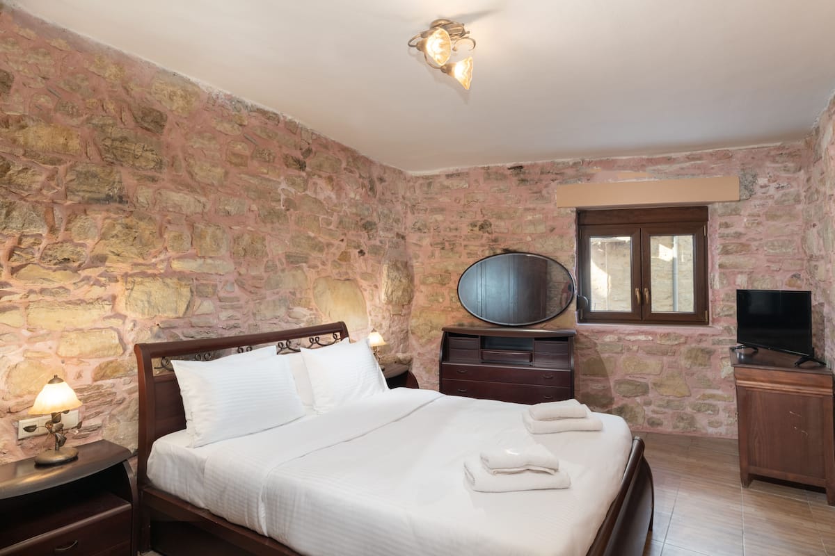 Een slaapkamer met een stenen muur, een bed met witte lakens, een nachtkastje met een lamp, een dressoir en een televisie.