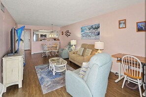 Ein Wohnzimmer mit Sofa, Sessel und einem Essbereich mit Tisch und Stühlen. Im Hintergrund ist eine Küche zu sehen.