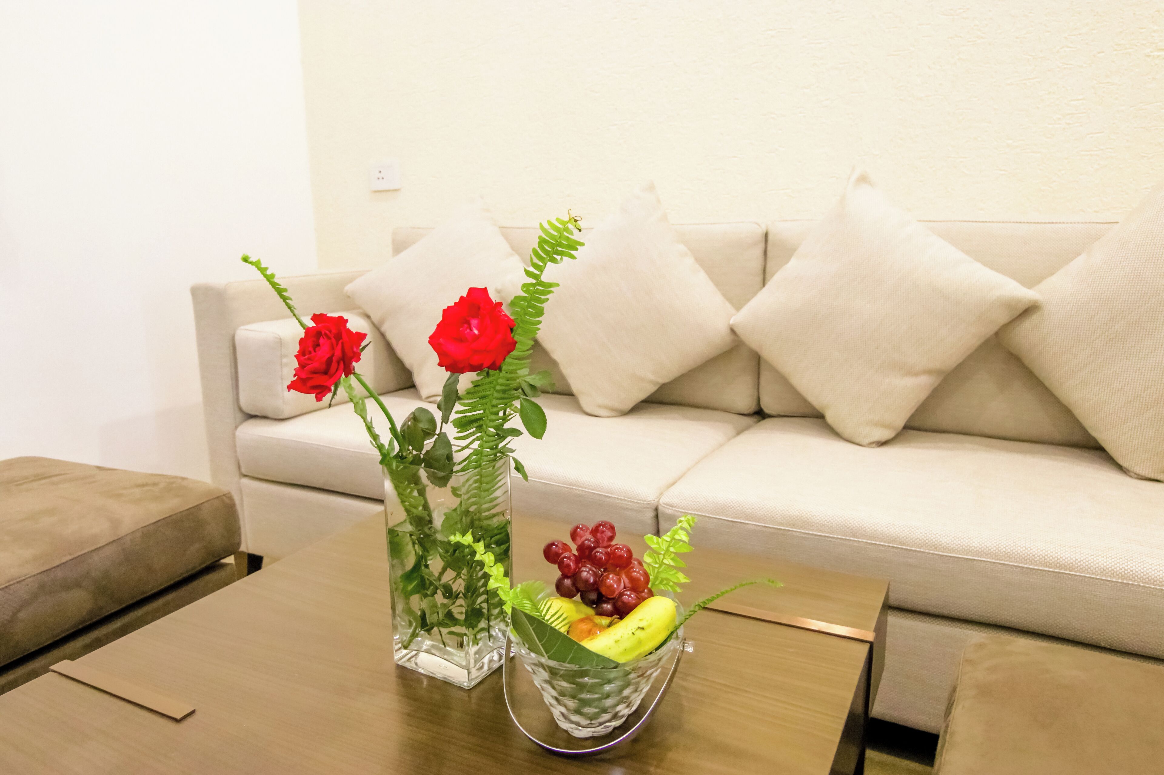 Ein Wohnzimmer mit beiger Couch, einem Holztisch und einer Glasvase mit roten Rosen sowie einer Obstschale.