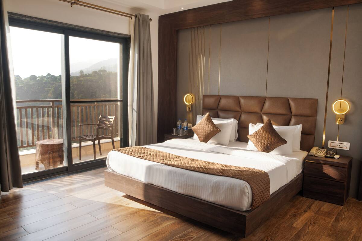 Une chambre spacieuse avec un grand lit, un plancher en bois, un balcon avec une chaise, et une table de chevet avec une lampe et un téléphone.