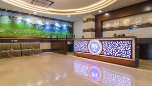 Uma área de recepção de hotel com design moderno, incluindo um grande mural, um balcão de recepção com visor digital e uma área de estar com cadeiras estofadas.
