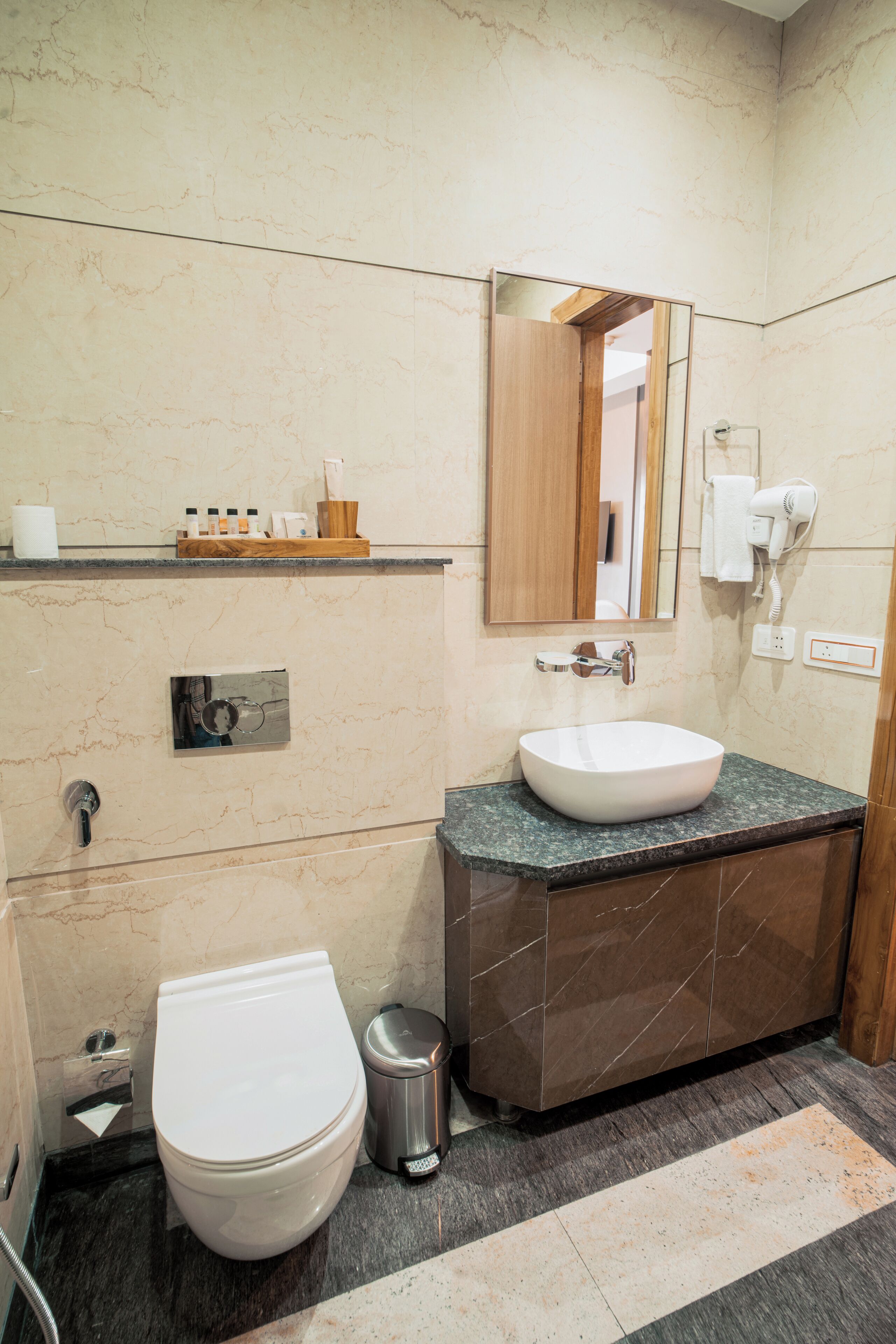 Un baño con inodoro blanco, un lavabo con encimera de mármol, un espejo y un secador de pelo en la pared.