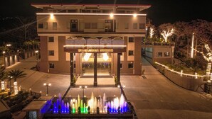 Entrada de hotel com uma fonte, um letreiro com a inscrição "DHAULADHAR HEIGHTS" e um caminho bem iluminado que leva à entrada.