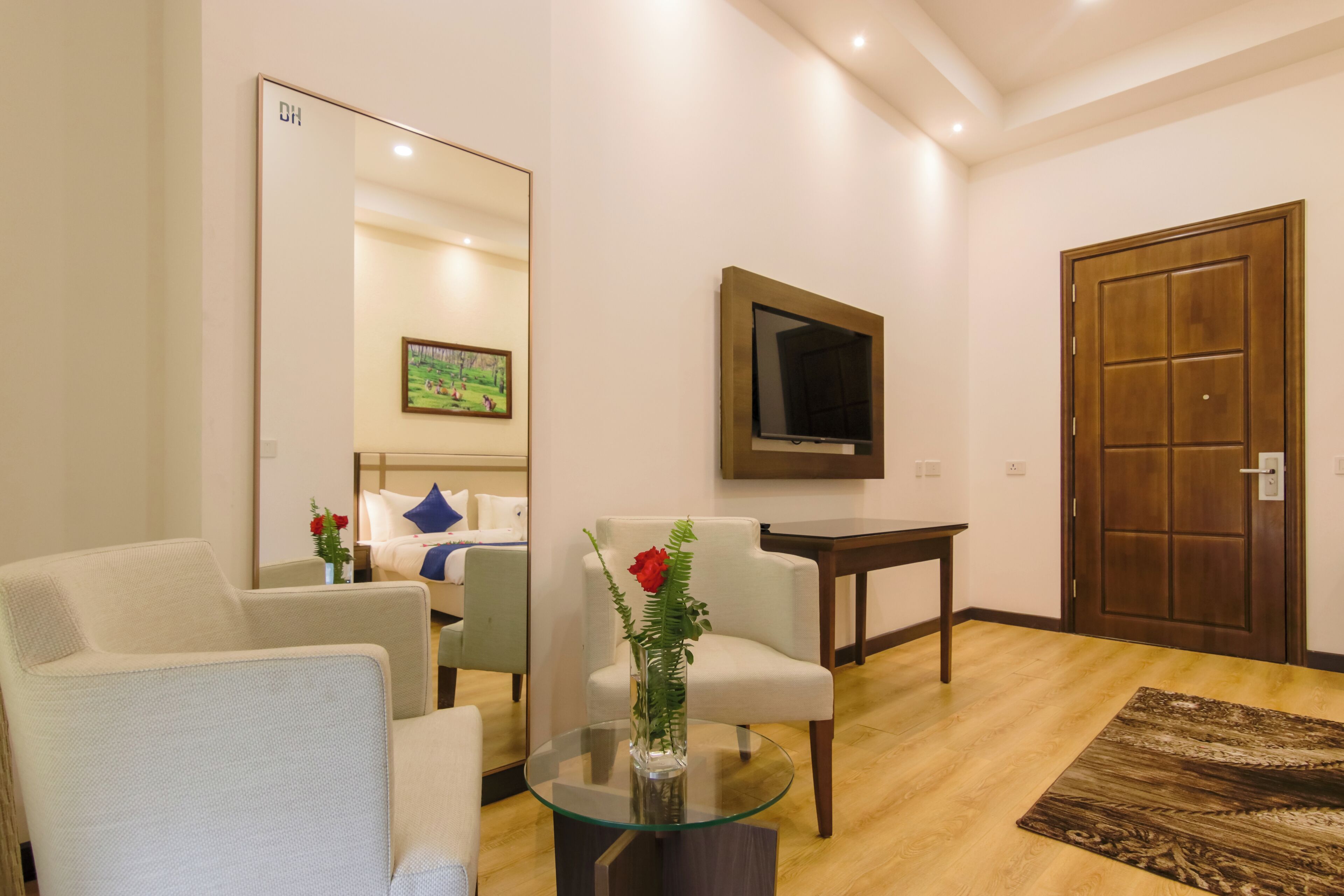 A modern living room with a glass coffee table, two white armchairs, a wooden door, a TV mounted on the wall, and a framed picture above the door.