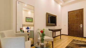 A modern living room with a glass coffee table, two white armchairs, a wooden door, a TV mounted on the wall, and a framed picture above the door.