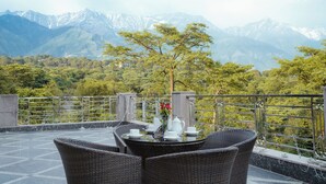 Um terraço com mobiliário de vime, uma mesa posta com chávenas brancas e um vaso de flores vermelhas, com montanhas ao fundo.