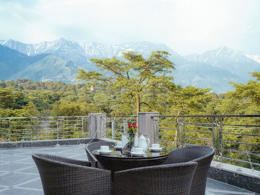 A terrace with wicker furniture, a table set with white cups and a vase of red flowers, and a mountain backdrop.
