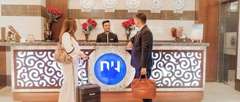 A hotel reception desk with a blue logo, clocks displaying times from various cities, and decorative vases with red flowers.