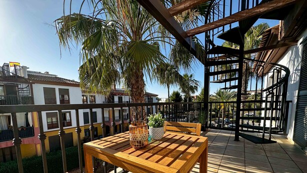 A wooden table and bench on a balcony with a view of neighboring buildings and a palm tree.