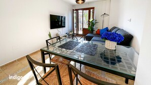 A modern living room with a glass-top dining table, black chairs, a sofa with blue and orange cushions, and a flat-screen TV mounted on the wall.