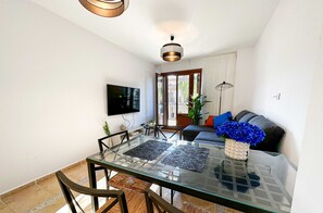 A modern living room with a glass-top dining table, black chairs, a sofa with blue and orange cushions, and a flat-screen TV mounted on the wall.