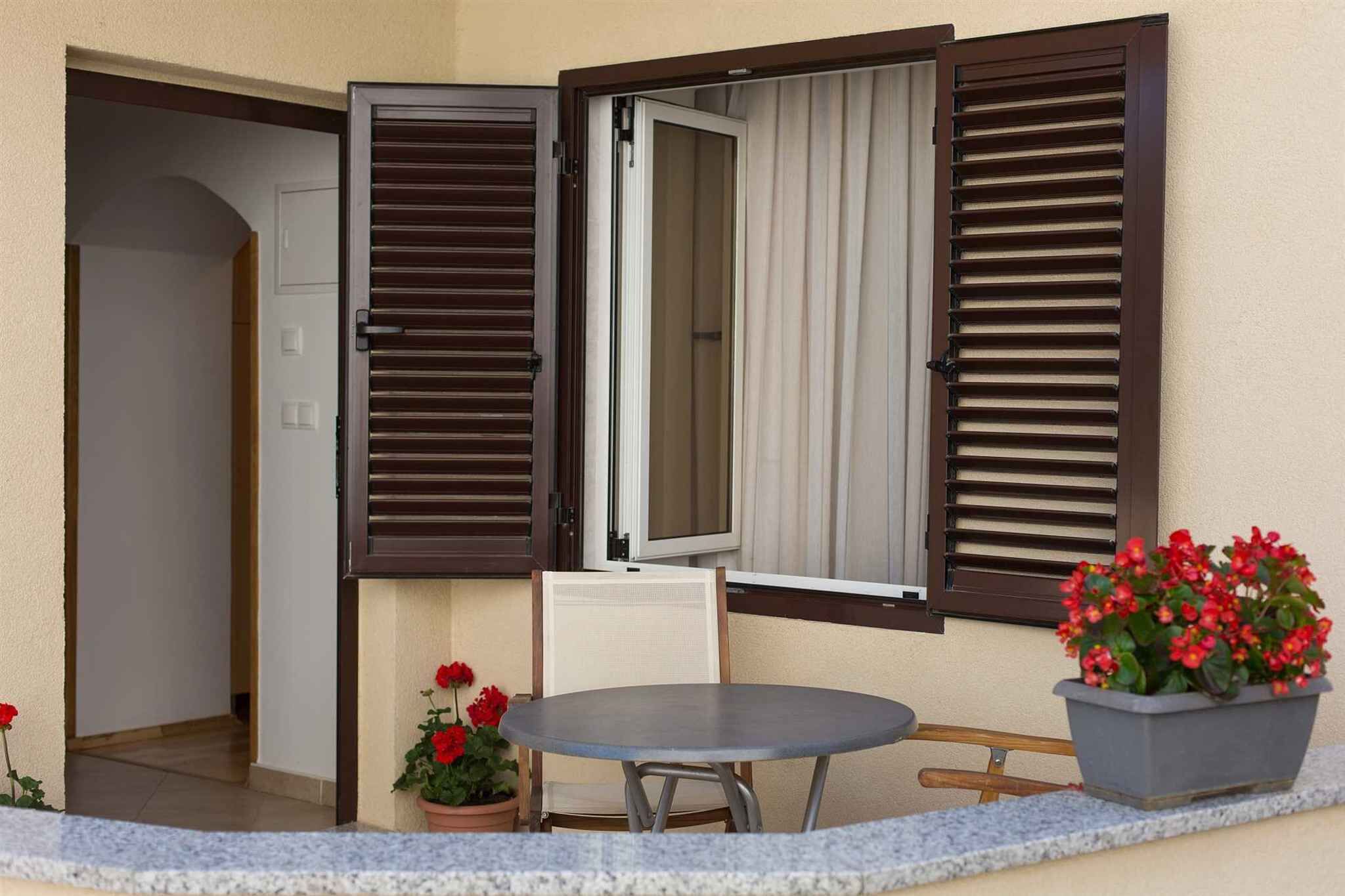 An outdoor seating area with a table and chairs, a potted plant with red flowers, and a window with brown shutters.