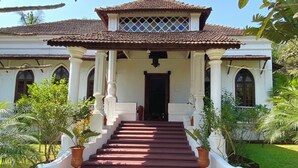 A two-story building with a tiled roof, a central entrance with steps, and a garden with potted plants. - VILLA LOU GOA Heritage Portuguese House built 1903 Exclusive Use of Private Pool (VERLA)