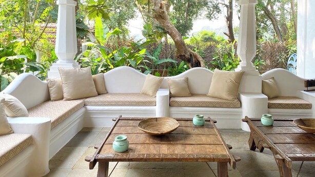 A covered seating area with a sofa and two wooden tables, surrounded by lush greenery. - VILLA LOU GOA Heritage Portuguese House built 1903 Exclusive Use of Private Pool (VERLA)