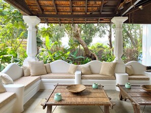 A covered seating area with a sofa and two wooden tables, surrounded by lush greenery. - VILLA LOU GOA Heritage Portuguese House built 1903 with Private Pool (VERLA)