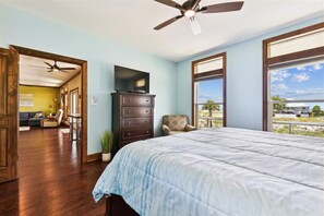 A bedroom with a large bed, a ceiling fan, a TV, and a view of the beach.
