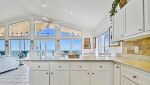 Une cuisine spacieuse avec des meubles blancs, un grand îlot central et de nombreuses fenêtres offrant une vue sur l’océan.