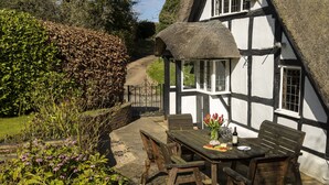 A traditional half-timbered house with a thatched roof, a wooden table with chairs, and a garden with purple flowers. - Corner Thatch - Worcestershire, a charming cottage sleeps 4 guests in 2 bedrooms (Abbots Morton)