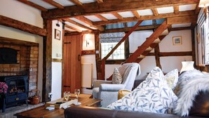 A cozy living room with exposed wooden beams, a fireplace, a comfortable sofa, and a dining table with a glass of wine. - Corner Thatch - Worcestershire, a charming cottage sleeps 4 guests in 2 bedrooms (Abbots Morton)