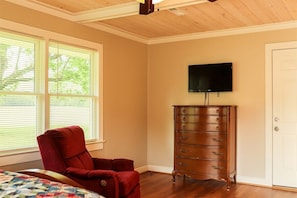 A bedroom with a wooden dresser, a red chair, a bed, a television on the wall, and a window with blinds.
