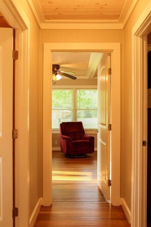 A hallway with a wooden floor leading to a room with a red chair and a ceiling fan.