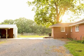 A stable with a covered area and a brick building, surrounded by a fenced area and trees.