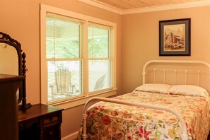 Un dormitorio con cama, ventana con persianas, un cuadro enmarcado en la pared y un aparador de madera.
