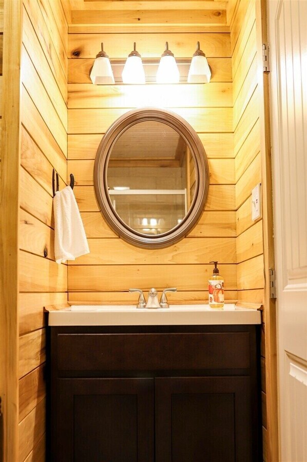 A wooden interior with a round mirror above a sink, three wall-mounted lights, and a towel hanging on a hook.