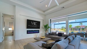Un amplio salón con un sofá grande de color gris, un televisor de pantalla plana montado en la pared y vistas al exterior a través de amplias ventanas.