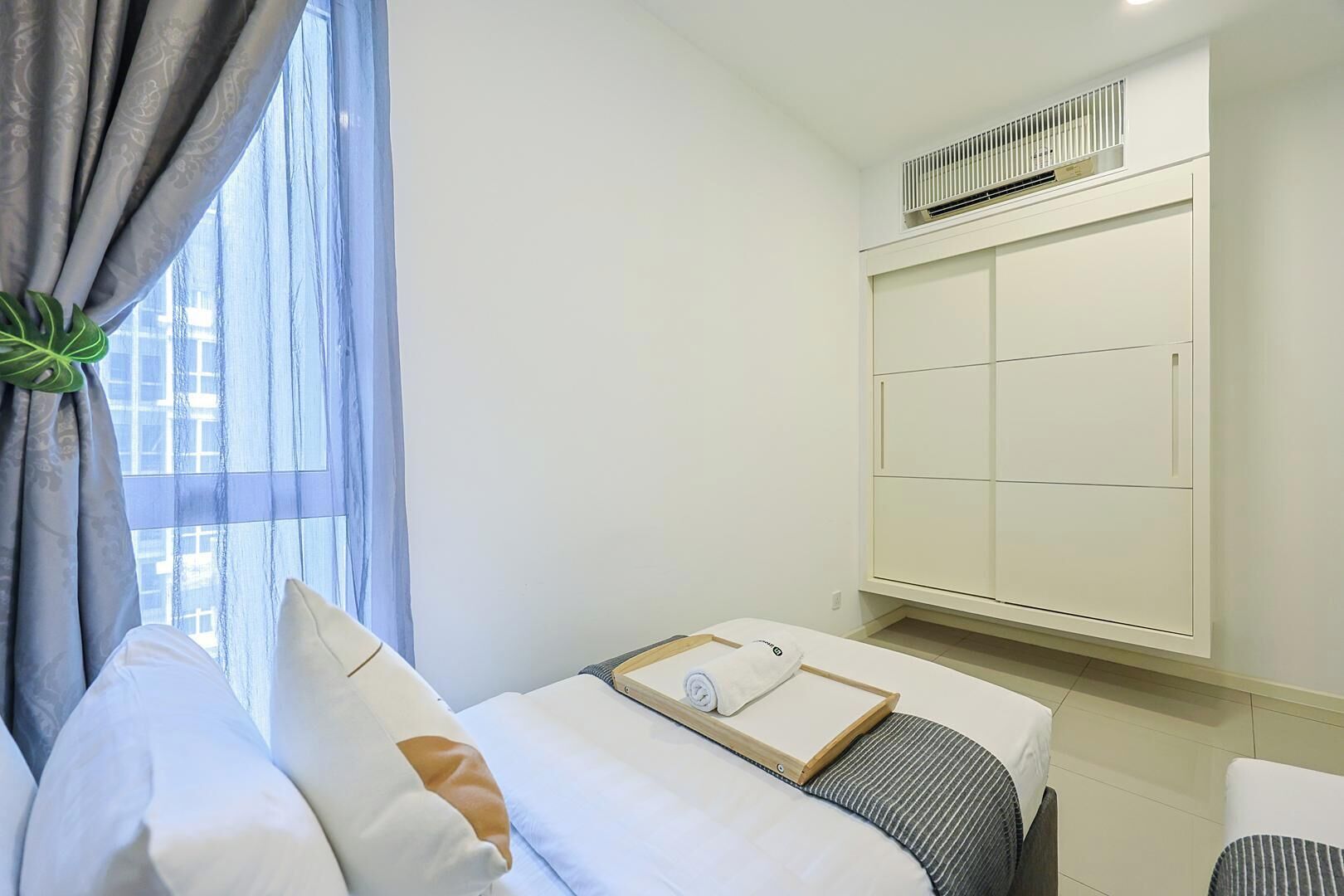 A hotel room with a bed, a bedside table, a window with curtains, and a wall-mounted air conditioning unit.